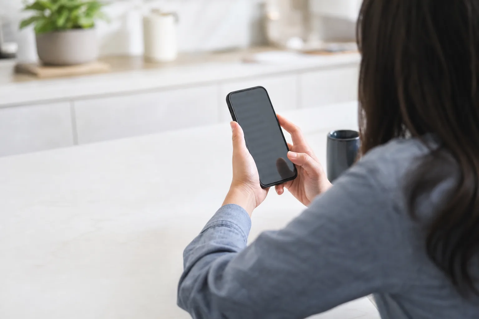 Over-the-shoulder view of a person reviewing a blurred finance app on a smartphone at a bright kitchen counter.